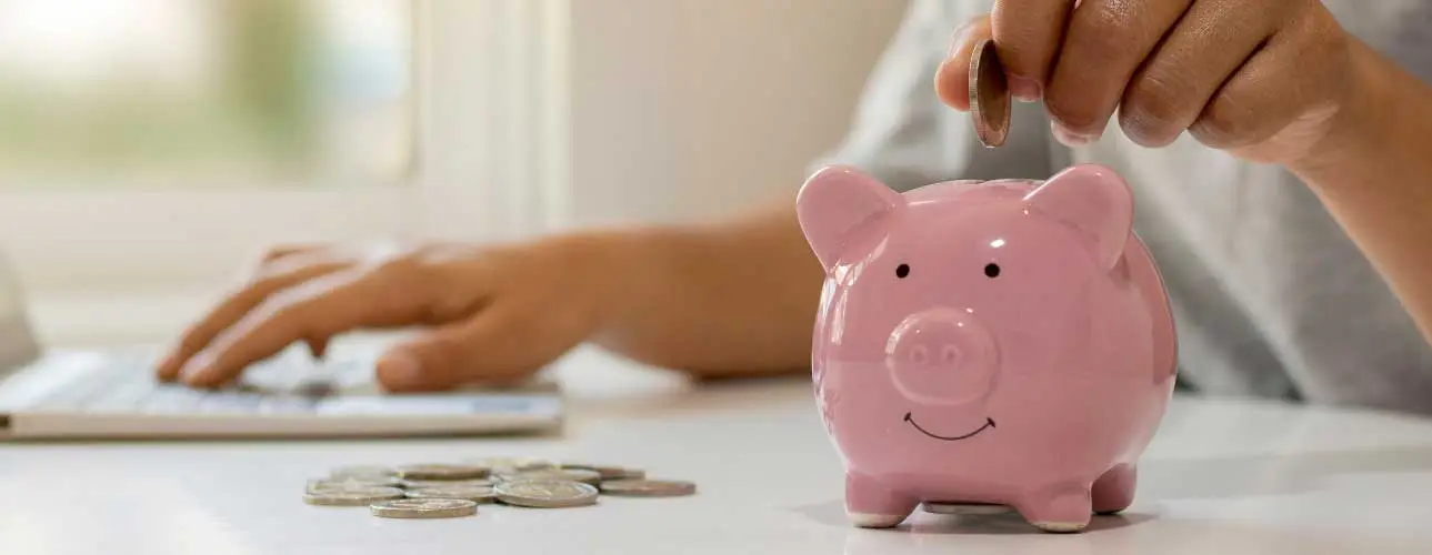 erson wirft Münze in rosafarbenes Sparschwein, im Hintergrund wird am Laptop gearbeitet – Symbol für Förderung & digitale Antragstellung.