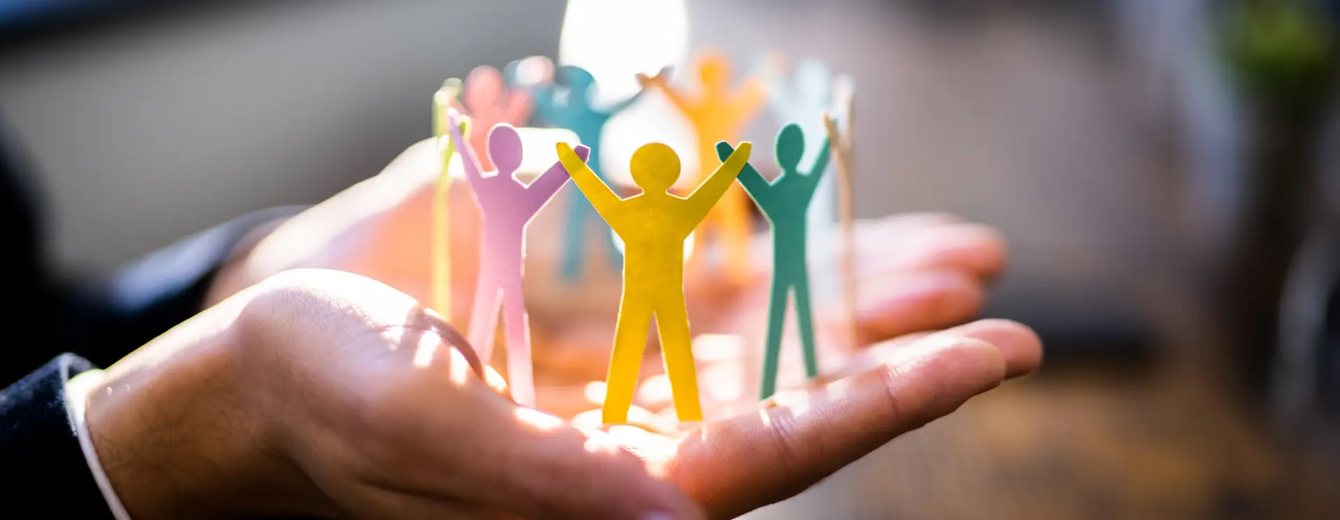 Symbolische Darstellung von Teamgeist – bunte Papierfiguren in einem Kreis auf schützenden Händen, im Lichtschein.