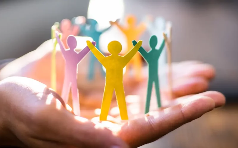 Symbolische Darstellung von Teamgeist – bunte Papierfiguren in einem Kreis auf schützenden Händen, im Lichtschein.